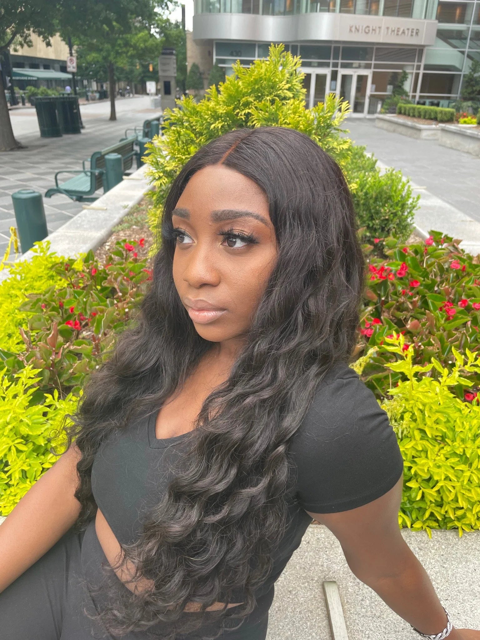 Woman with long, wavy hair sitting outdoors in front of greenery and a building wearing a custom loose wave wig from WinkLilly beauty.